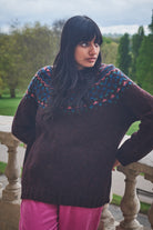 Woman wearing a dark sweater with colorful patterns, standing on a stone balcony with greenery in the background.