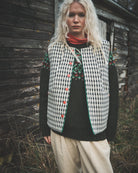 A woman stands outdoors in front of a weathered wooden wall, wearing a black-and-white gingham vest with red buttons and embroidered green floral details on the chest, layered over a dark sweater and light beige pants.