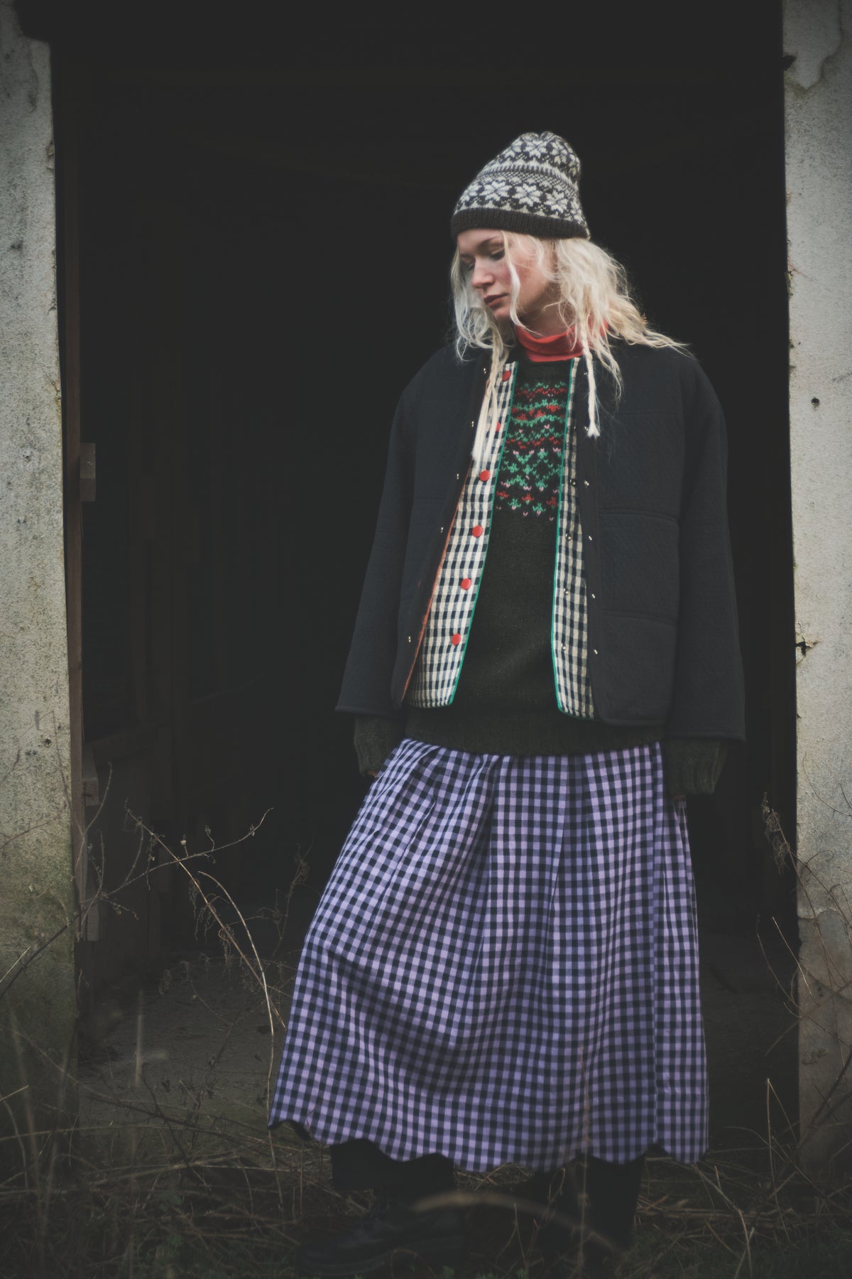 A person standing in a dark doorway wearing a black jacket, a patterned knit beanie, a red and green patterned sweater, and a blue-and-white gingham skirt.
