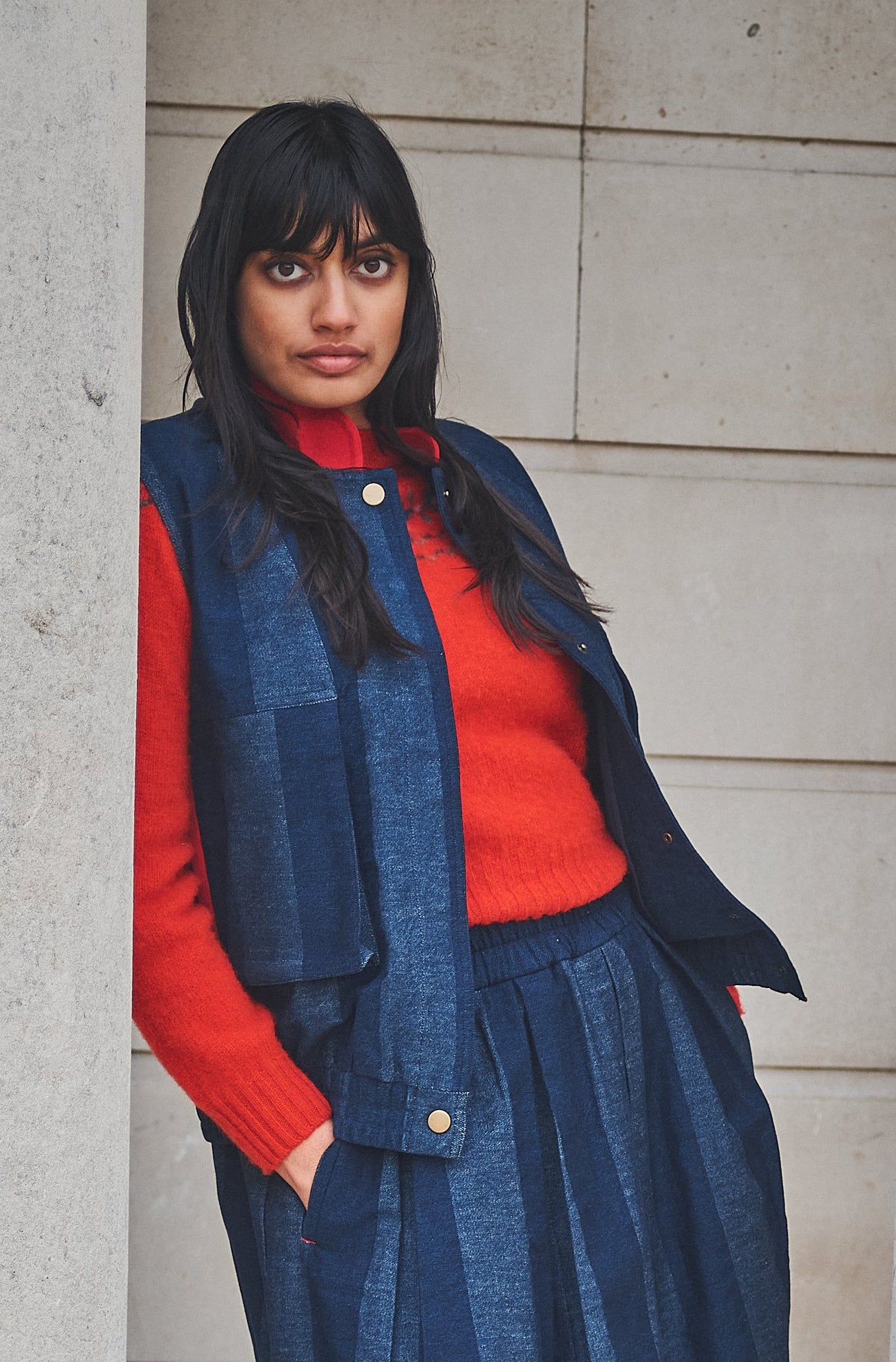 Woman in a Lowie denim stripe gilet, red fair isle jumper and blue trousers standing between stone columns.