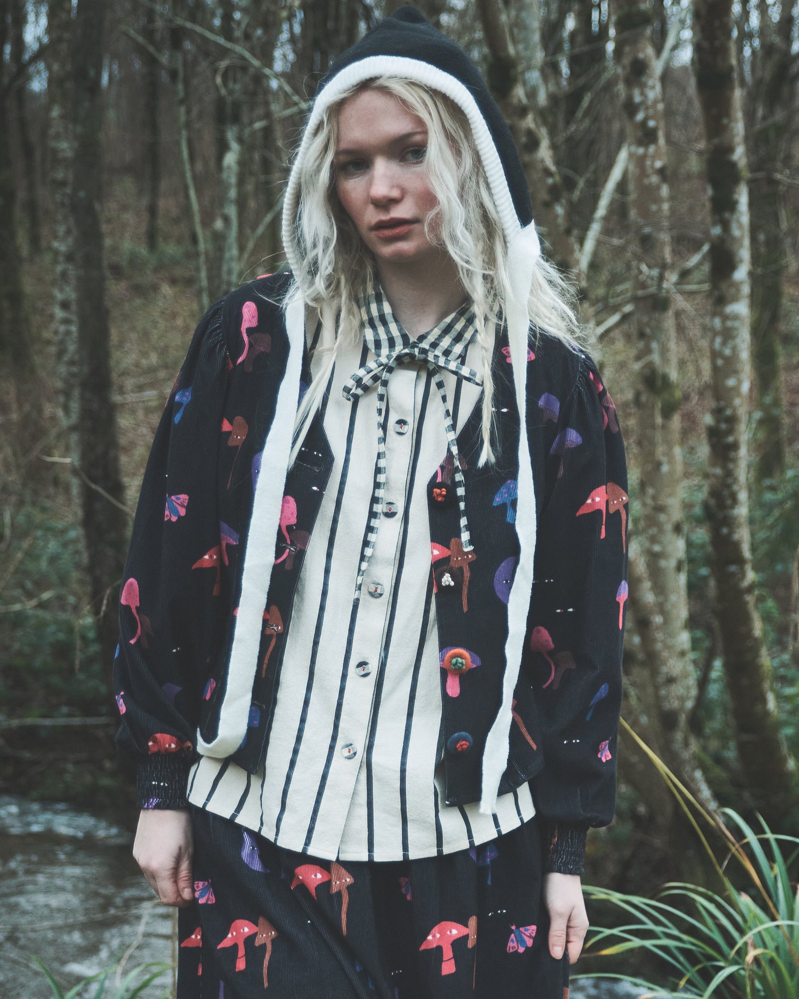 A model wearing a black mushroom blouse jacket, layered over a cream and black striped shirt, standing in a forest.