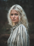 A woman with wavy platinum blonde hair wearing a cream-colored blouse with thin black vertical stripes and a matching striped necktie, photographed outdoors against a dark, blurred forest background.