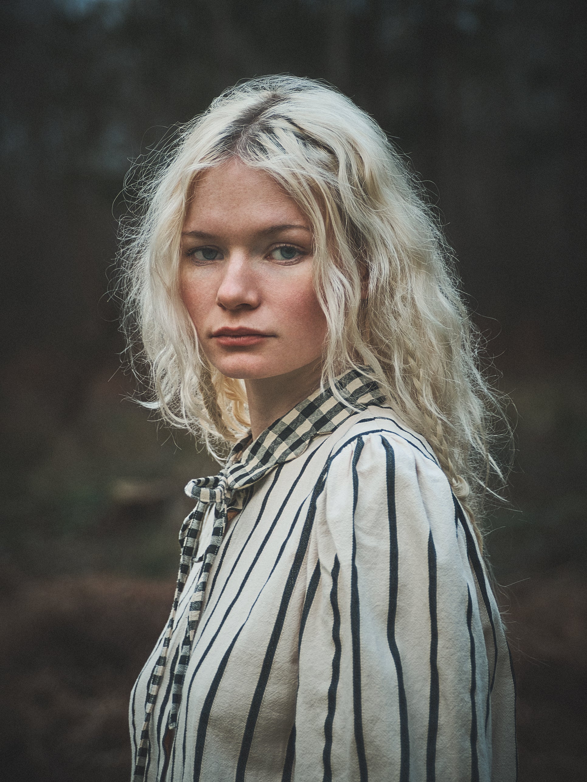 A woman with wavy platinum blonde hair wearing a cream-colored blouse with thin black vertical stripes and a matching striped necktie, photographed outdoors against a dark, blurred forest background.