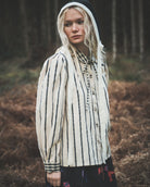 A woman wearing a cream-colored cotton shirt with black vertical stripes, a black knit hood, and a dark skirt stands in a forest clearing.