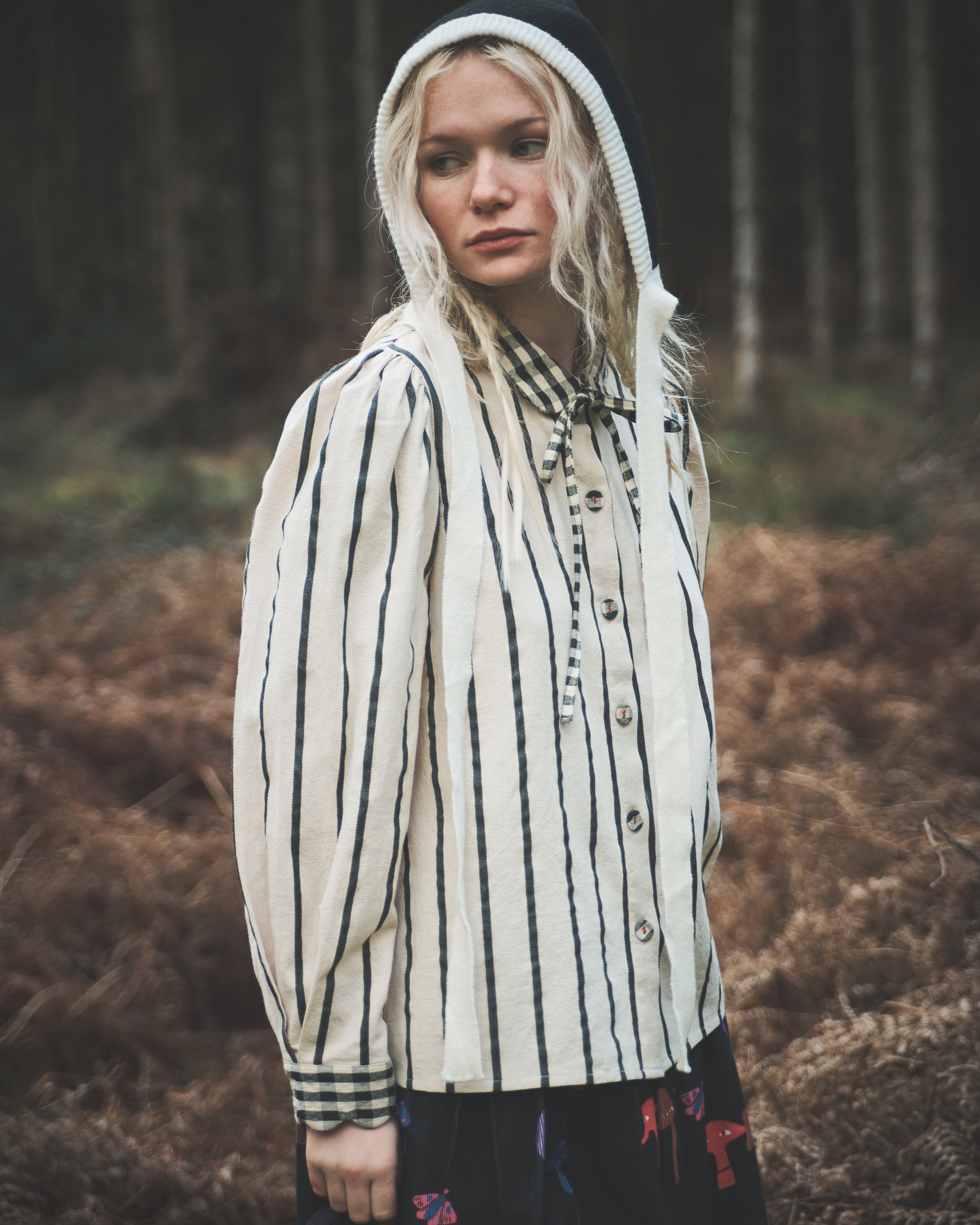 A woman wearing a cream-colored cotton shirt with black vertical stripes, a black knit hood, and a dark skirt stands in a forest clearing.