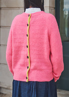 Person wearing a pink cardigan with a yellow trim and black buttons, standing against a neutral background.