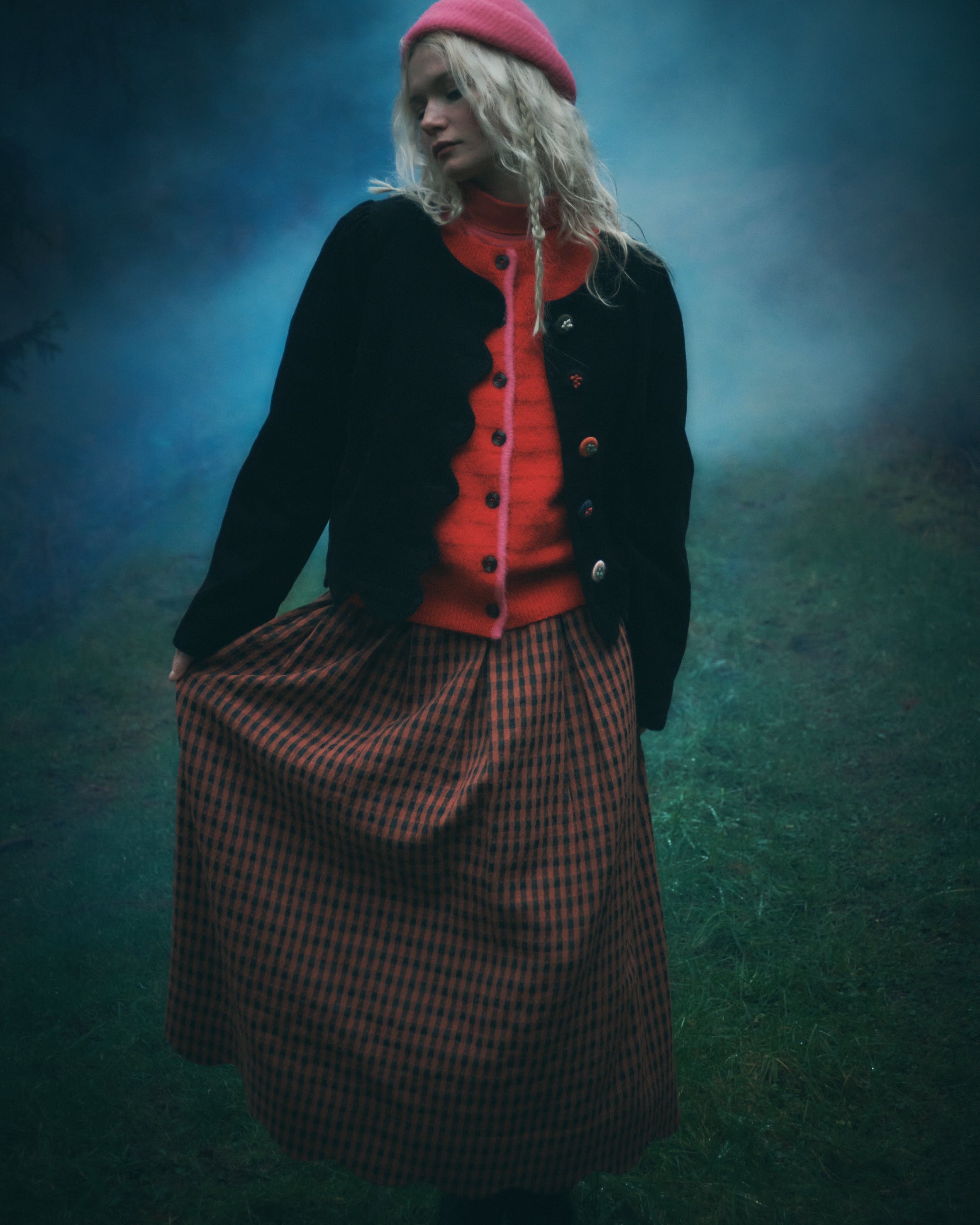 A woman stands outdoors in a misty blue-green environment wearing a bright pink beret, a black cardigan over a vibrant orange-red sweater, and a brown and black checkered skirt.