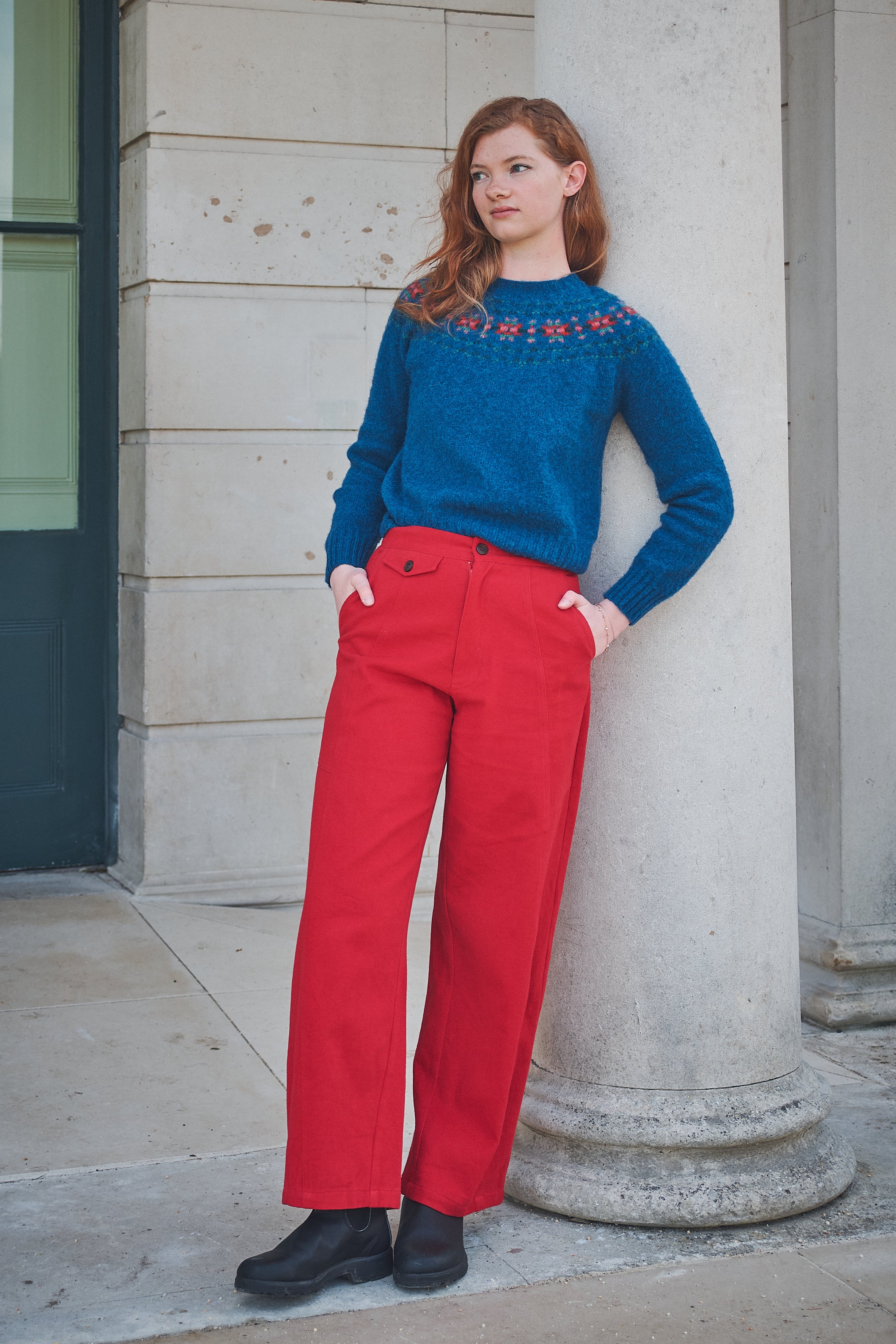 Woman wearing a blue sweater with red patterns and red pants standing against a stone building.