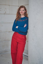 Woman wearing a blue sweater with red patterns and red pants standing against a light-colored wall.