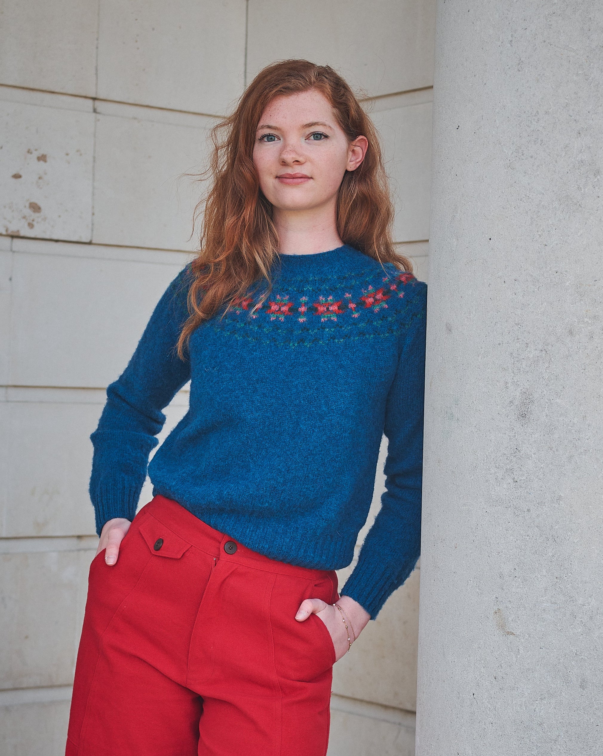 Woman wearing a blue sweater with red patterns and red pants standing against a light-colored wall.