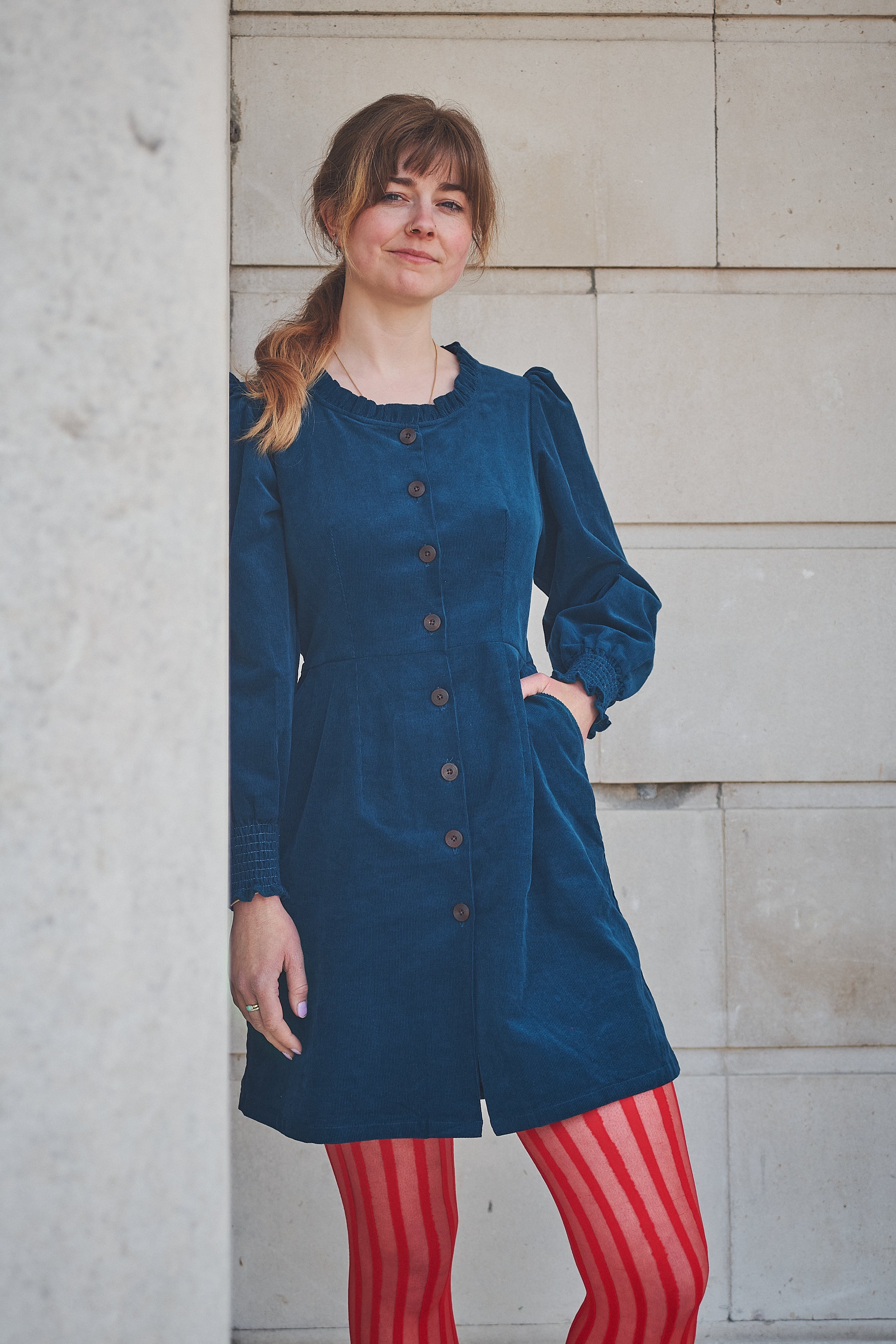 Woman wearing a blue dress with a button-up front, standing against a light stone wall.