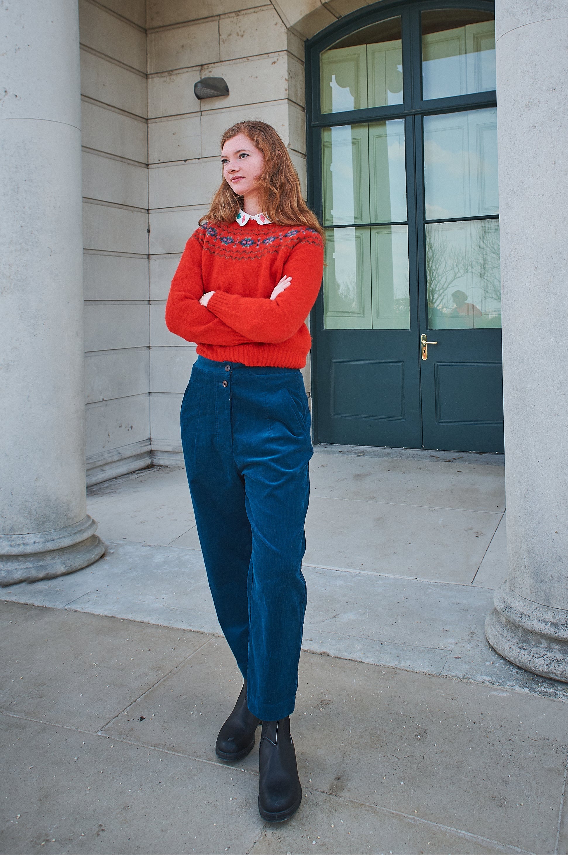 Person wearing a red sweater and blue pants standing in front of stone columns and a window.