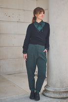 Woman wearing a dark sweater and green plaid pants standing against a stone wall.