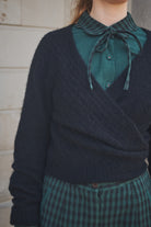 Person wearing a dark sweater over a green shirt with a bow tie, standing against a neutral background.