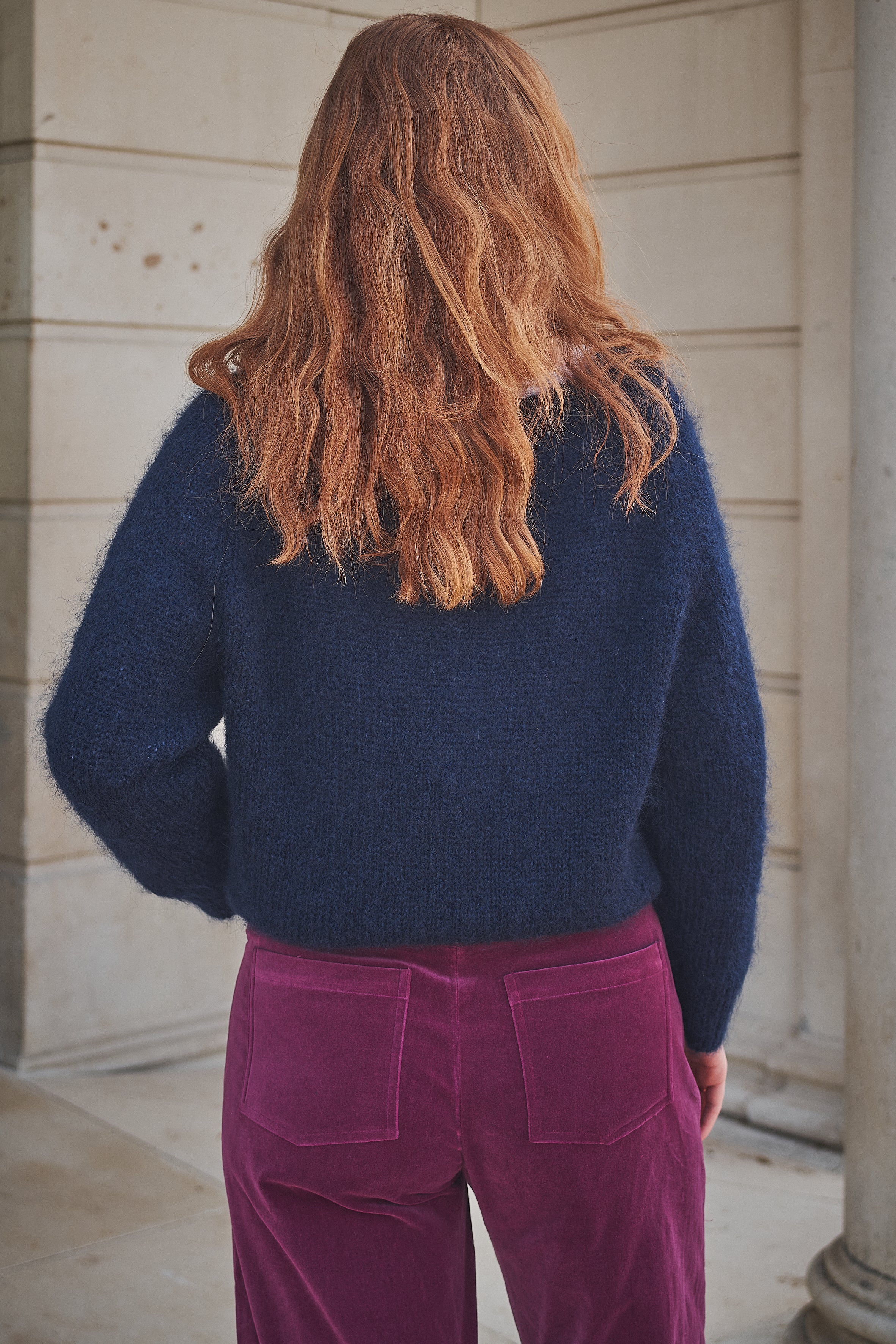 Person wearing a navy sweater and pink pants standing against a stone wall.