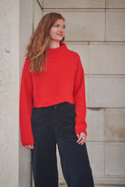 Woman wearing a red sweater and dark pants standing against a stone wall.