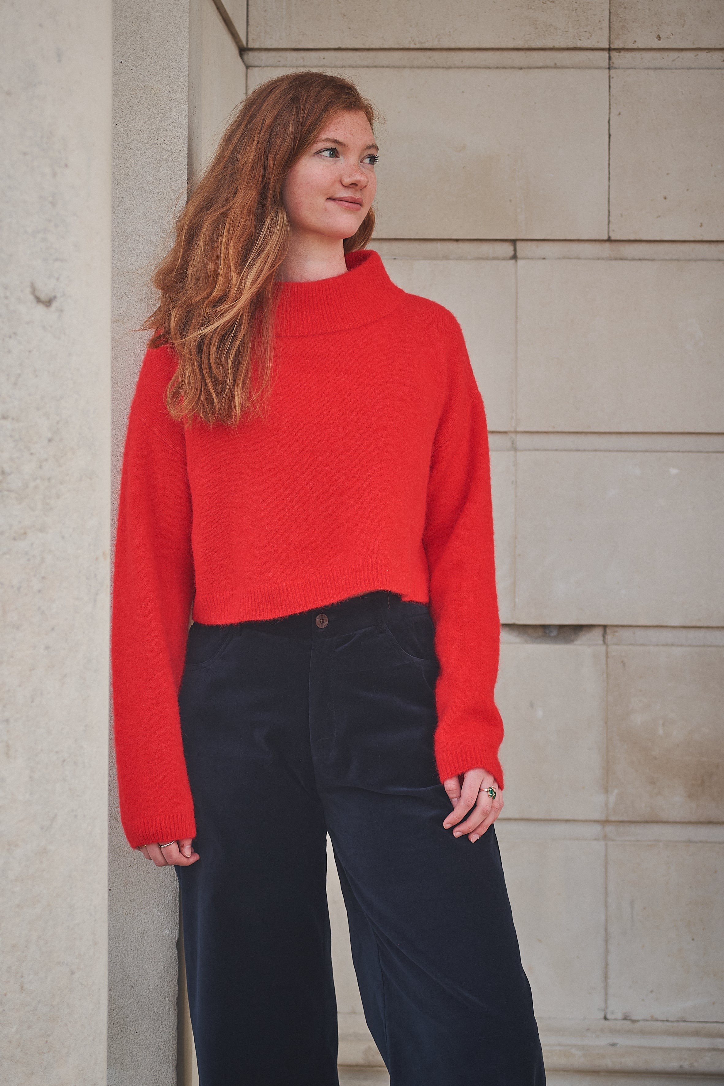 Woman wearing a red sweater and dark pants standing against a stone wall.