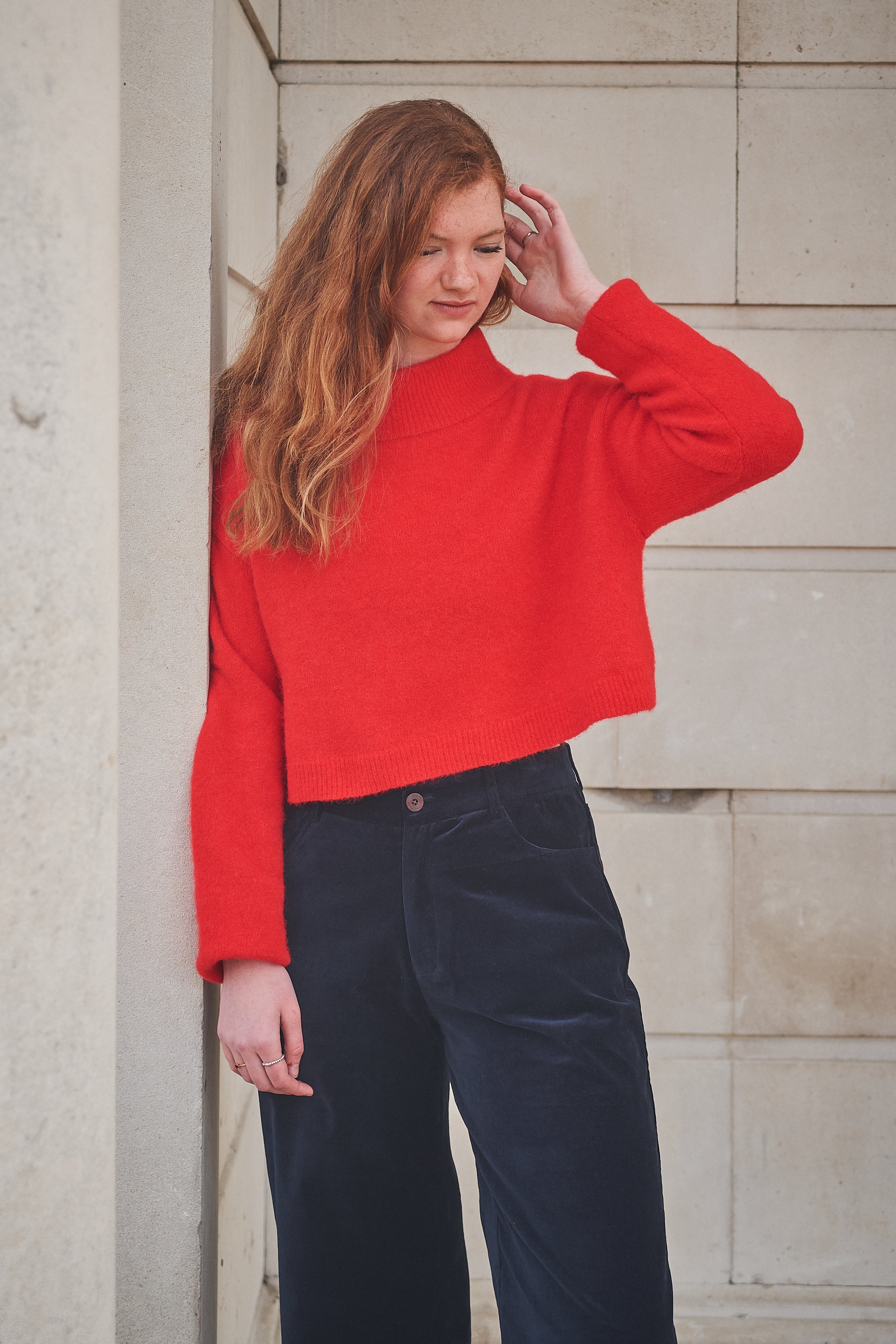 Woman wearing a red sweater and dark pants against a stone wall.
