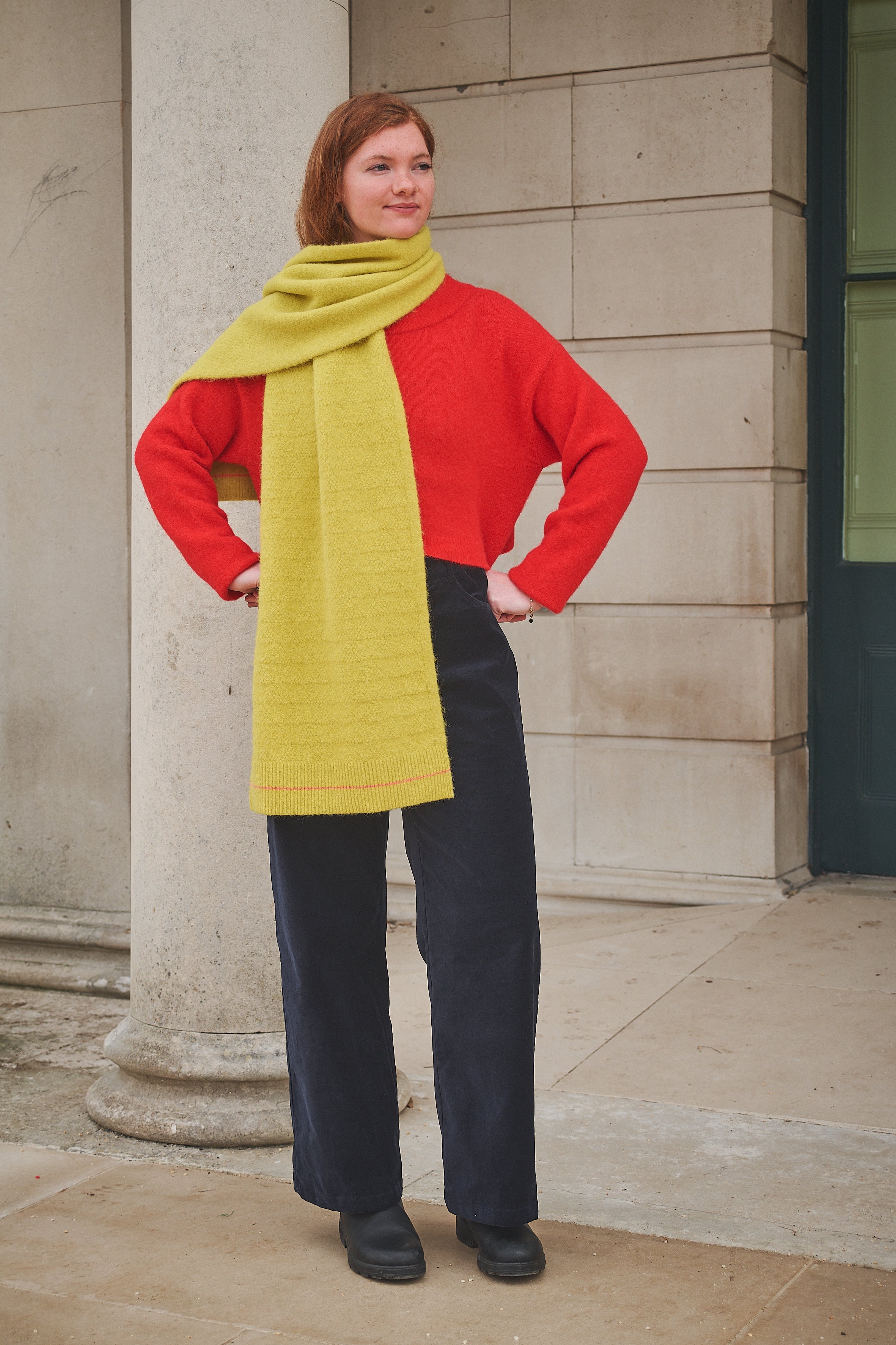 Woman wearing a red sweater, yellow scarf, and black pants standing against a stone building.