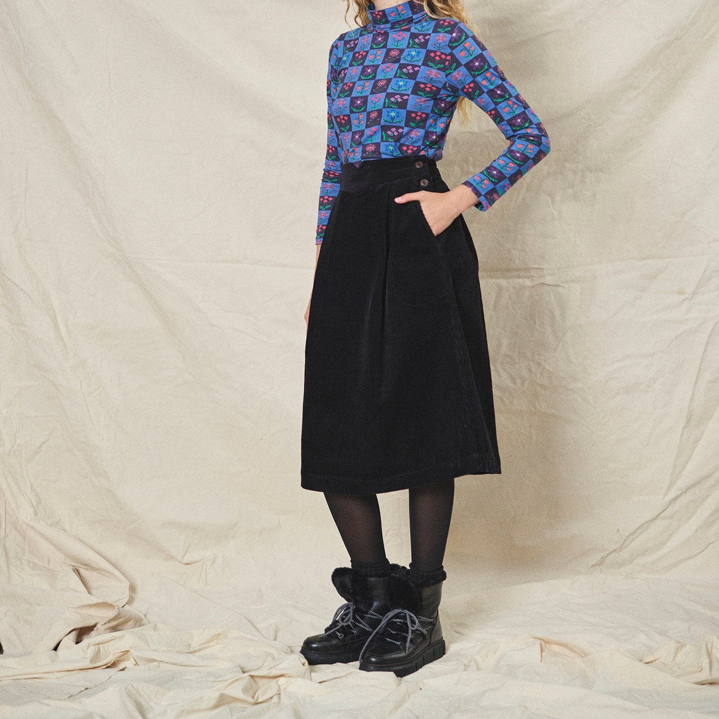 A person wearing a straight black corduroy skirt with a shaped waistband and deep inverted pleats, paired with a multicolored floral top and black boots.