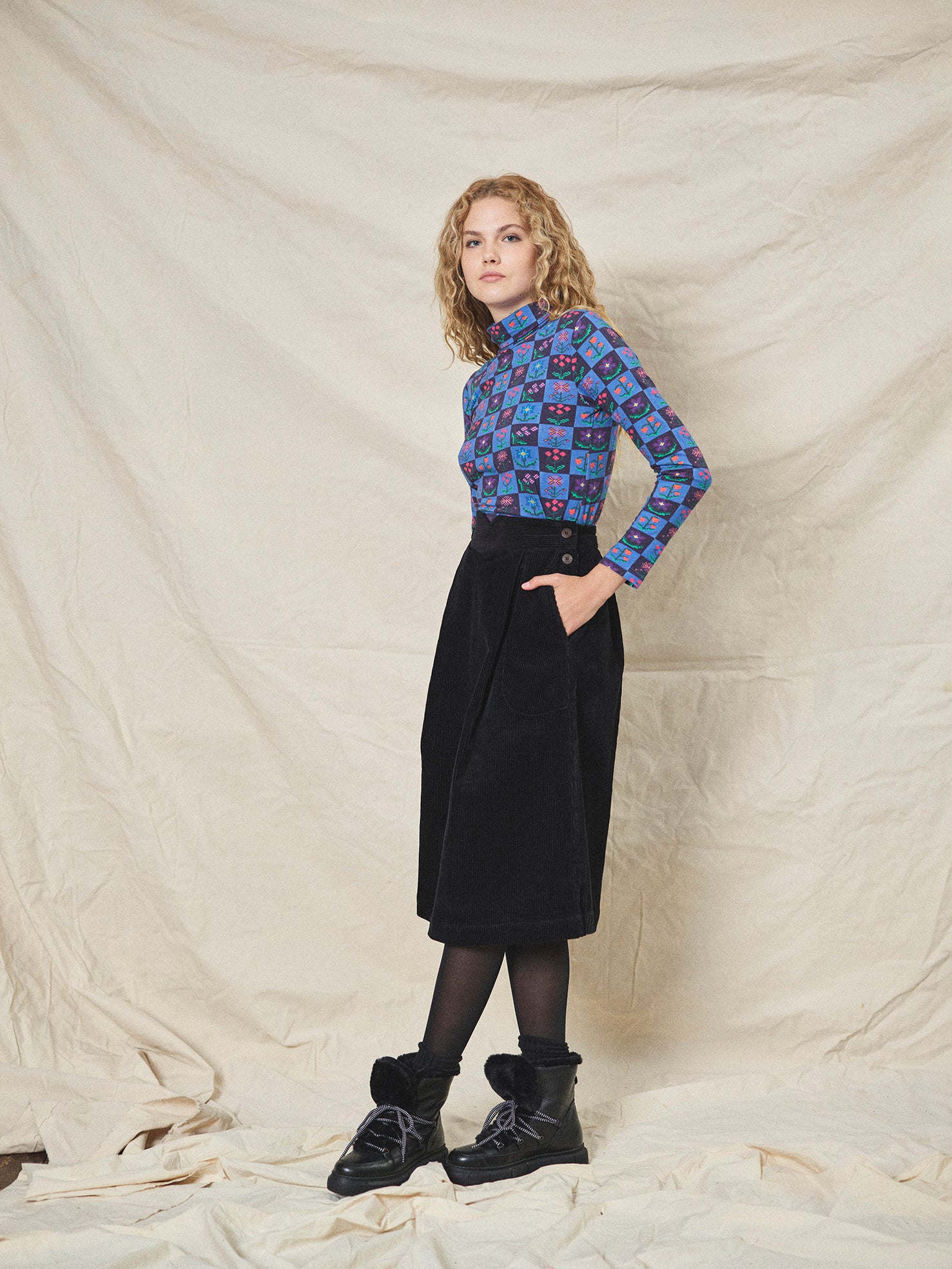 A person wearing a straight black corduroy skirt with a shaped waistband and deep inverted pleats, paired with a multicolored floral top and black boots.