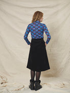 A person wearing a straight black corduroy skirt with a shaped waistband and deep inverted pleats, paired with a multicolored floral top and black boots.