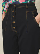 A pair of black cotton drill trousers with a high waist, wide beltband, and 5 button fly front. Features include slanted side pockets and raw stitching.