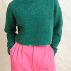 A model wearing bright pink wide leg trousers with pleats and a tab front opening, paired with a green sweater and brown boots.