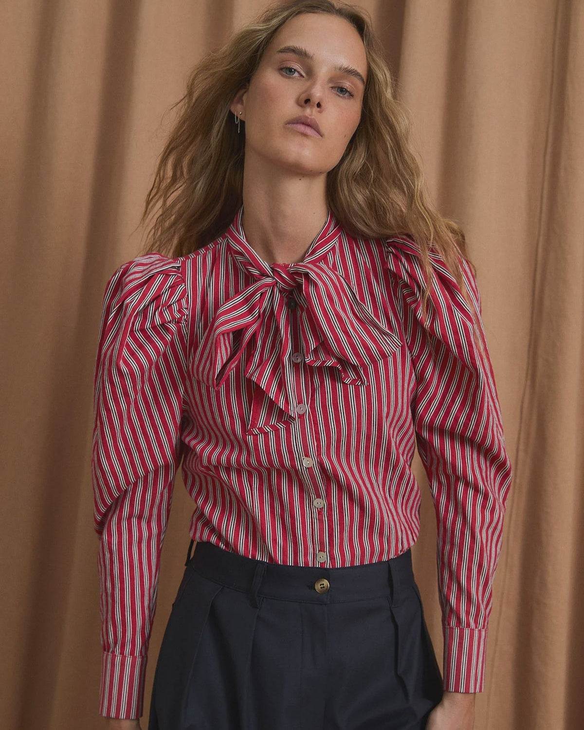 A woman wearing a red and white vertically striped long-sleeve blouse with ruffled details and a bow at the collar, paired with dark navy trousers, standing in front of a draped tan curtain.