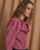A woman wearing a red and white vertically striped blouse with a large ruffled bow at the collar, paired with dark trousers, posed against a draped tan fabric backdrop.
