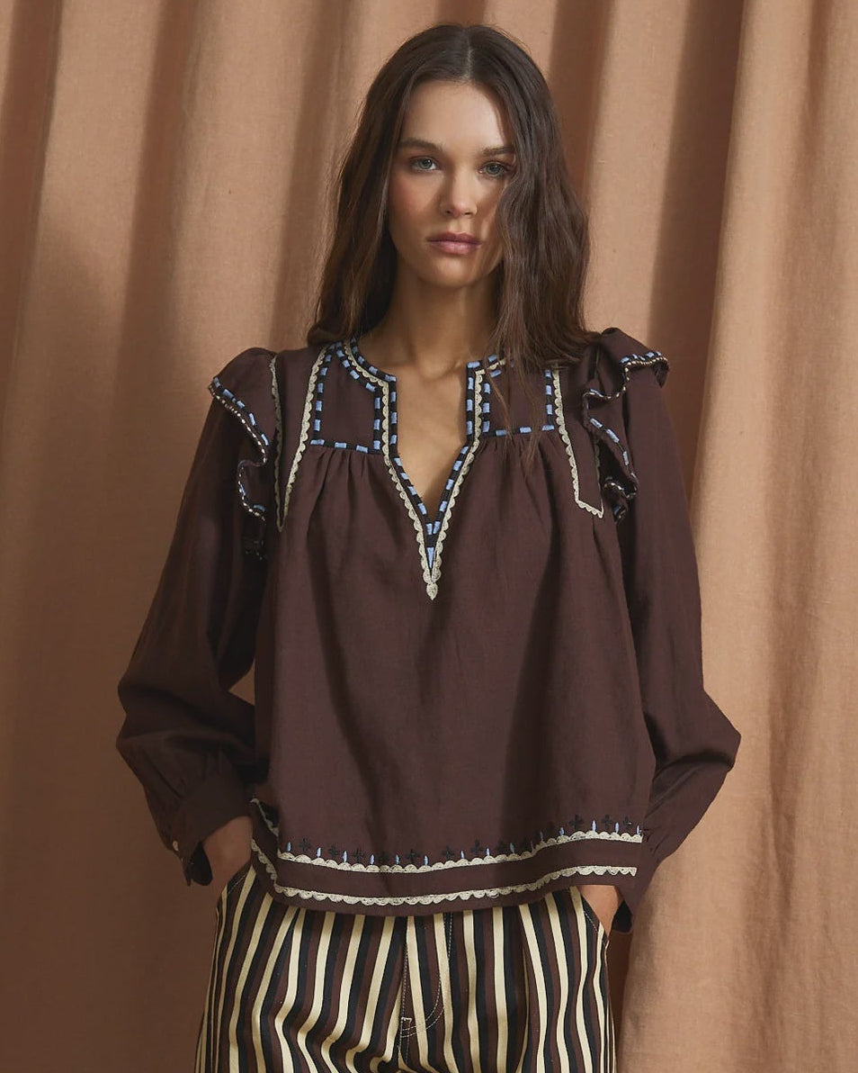 A woman modeling a dark brown long-sleeve blouse with embroidered detailing around the neckline and a striped black and cream wide-leg pant set, posed against a draped tan fabric backdrop.