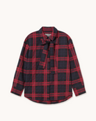 A long-sleeve button-up shirt with a red and black plaid pattern, featuring a self-tie neck bow and a small chest pocket, displayed flat against a light cream background.