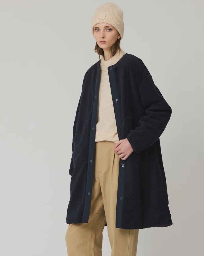 A model stands against a light gray studio background wearing a long navy cardigan over a cream turtleneck, beige wide-leg trousers, a beige beanie, and dark brown loafers.