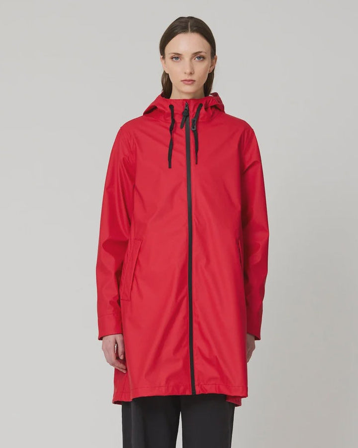 A model stands against a light gray studio background wearing a bright red hooded raincoat with a black front zipper and black drawstrings, paired with black pants and blue sneakers.