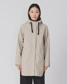A model stands against a light gray studio background wearing a light beige hooded raincoat with black drawstrings, paired with black wide-leg trousers and dark brown shoes.