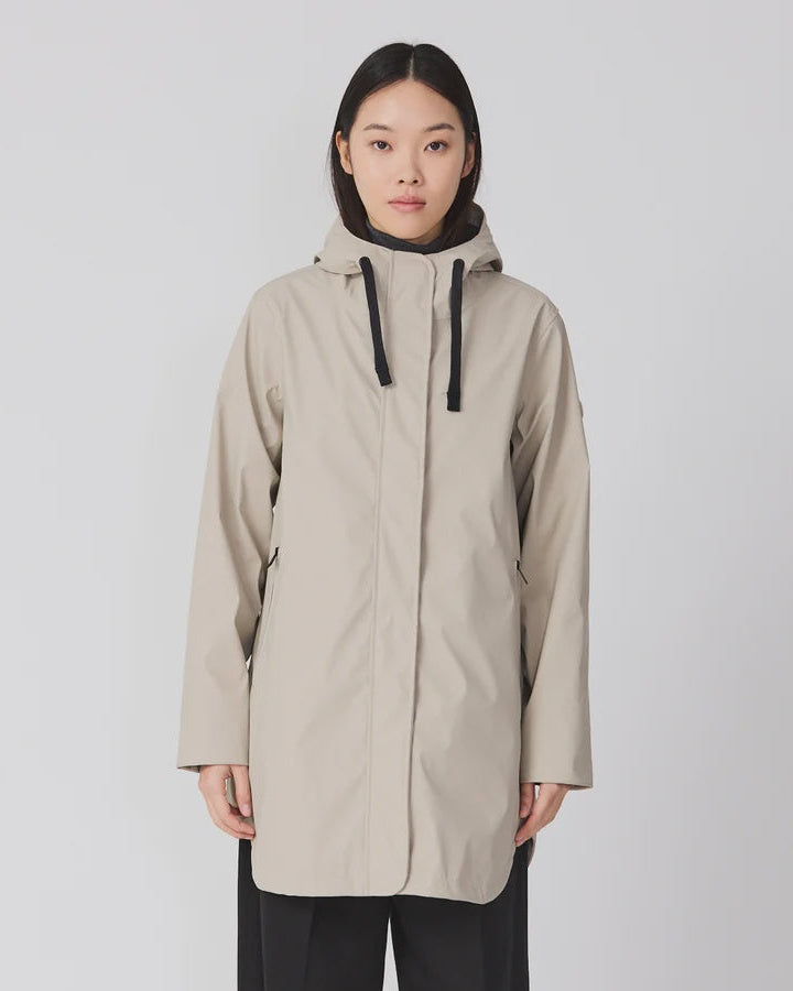 A model stands against a light gray studio background wearing a light beige hooded raincoat with black drawstrings, paired with black wide-leg trousers and dark brown shoes.