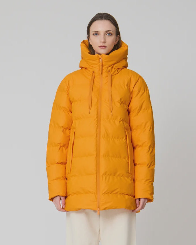 A woman stands against a light gray studio background wearing a bright mustard-yellow hooded puffer jacket, cream wide-leg pants, and white sneakers.