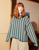 A woman with red hair wearing a green and white vertically striped cropped sweater and beige wide-leg pants, standing in a warm-toned indoor room.