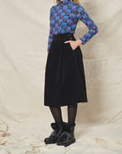 A person wearing a straight black corduroy skirt with a shaped waistband and deep inverted pleats, paired with a multicolored floral top and black boots.