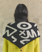 Person wearing a black and white patterned cape over a yellow garment against a beige wall.