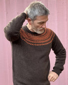 A man wearing a chocolate brown and orange fairisle pattern jumper with a ribbed round neck and long sleeves.