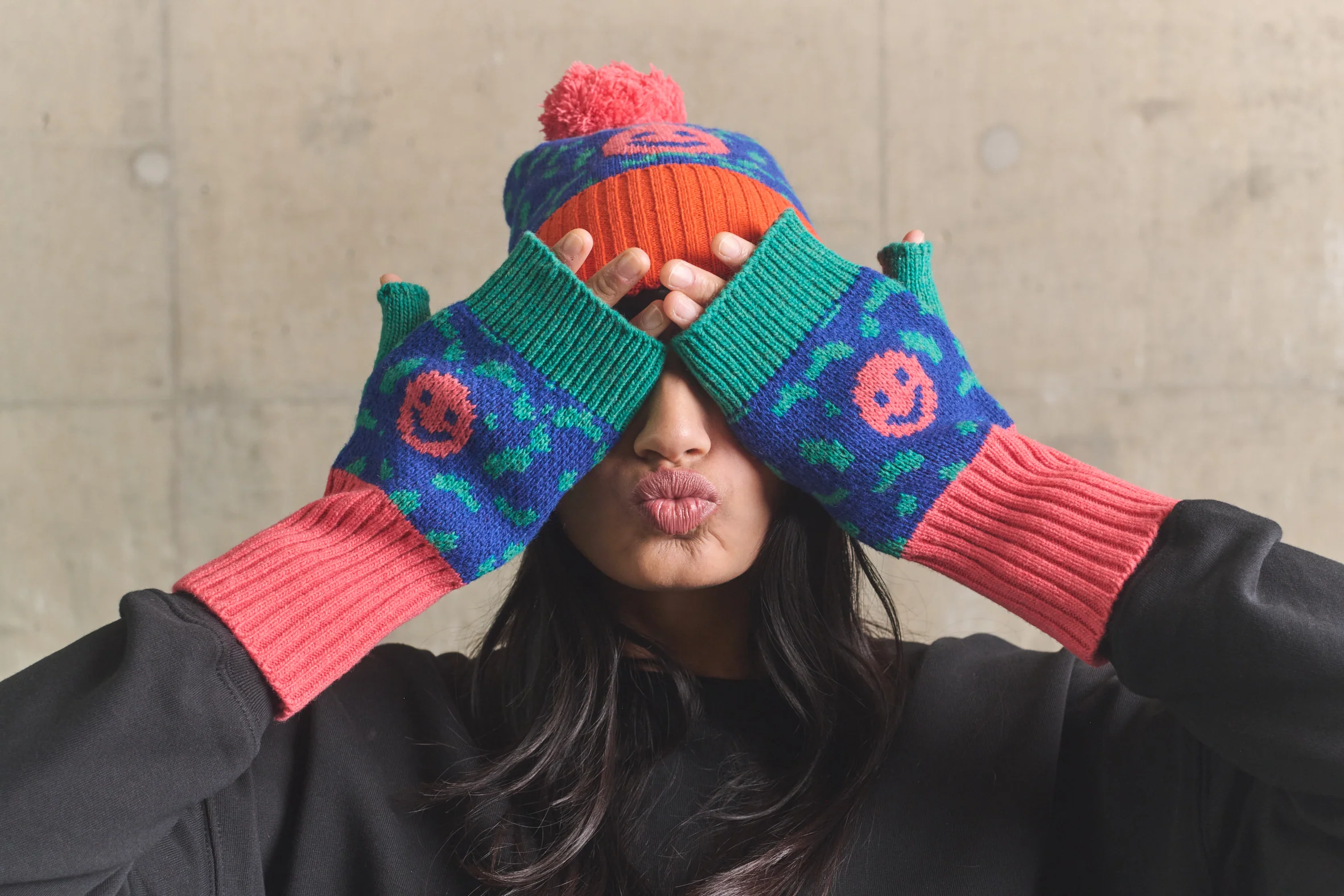 Person wearing colorful knitted hat and gloves against a concrete wall.