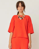 A woman wearing a vibrant orange, loose-fitting short-sleeved blouse with a round collar and side slits.