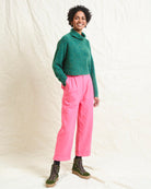 A model wearing bright pink wide leg trousers with pleats and a tab front opening, paired with a green sweater and brown boots.
