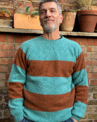 A man standing in front of a brick wall and potted plants, wearing a striped jumper with horizontal block colors, predominantly teal and brown, with a ribbed-knit hemline and ribbed trim around the neck and wrists.