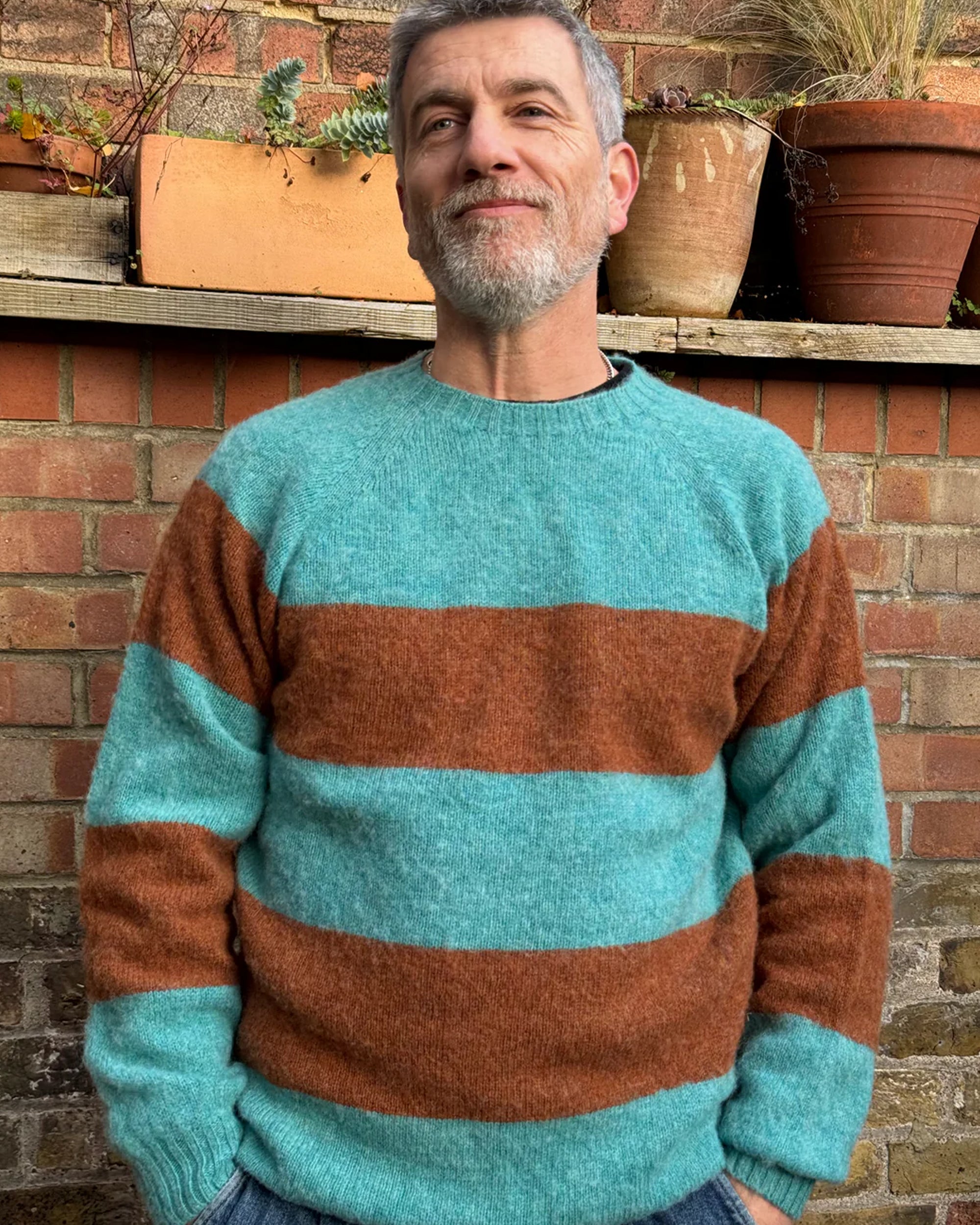 A man standing in front of a brick wall and potted plants, wearing a striped jumper with horizontal block colors, predominantly teal and brown, with a ribbed-knit hemline and ribbed trim around the neck and wrists.